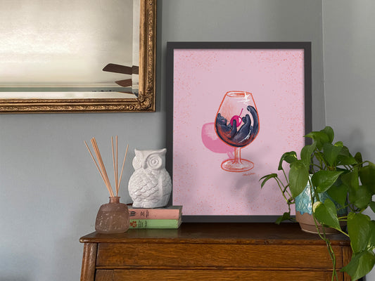 Framed art print of a black cat in a wine glass with a pink background on a wooden bookshelf.